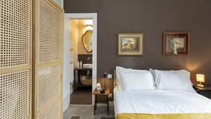 Un dormitorio con cama, mesitas de noche, lámpara, un cuadro enmarco y una puerta que da al baño.