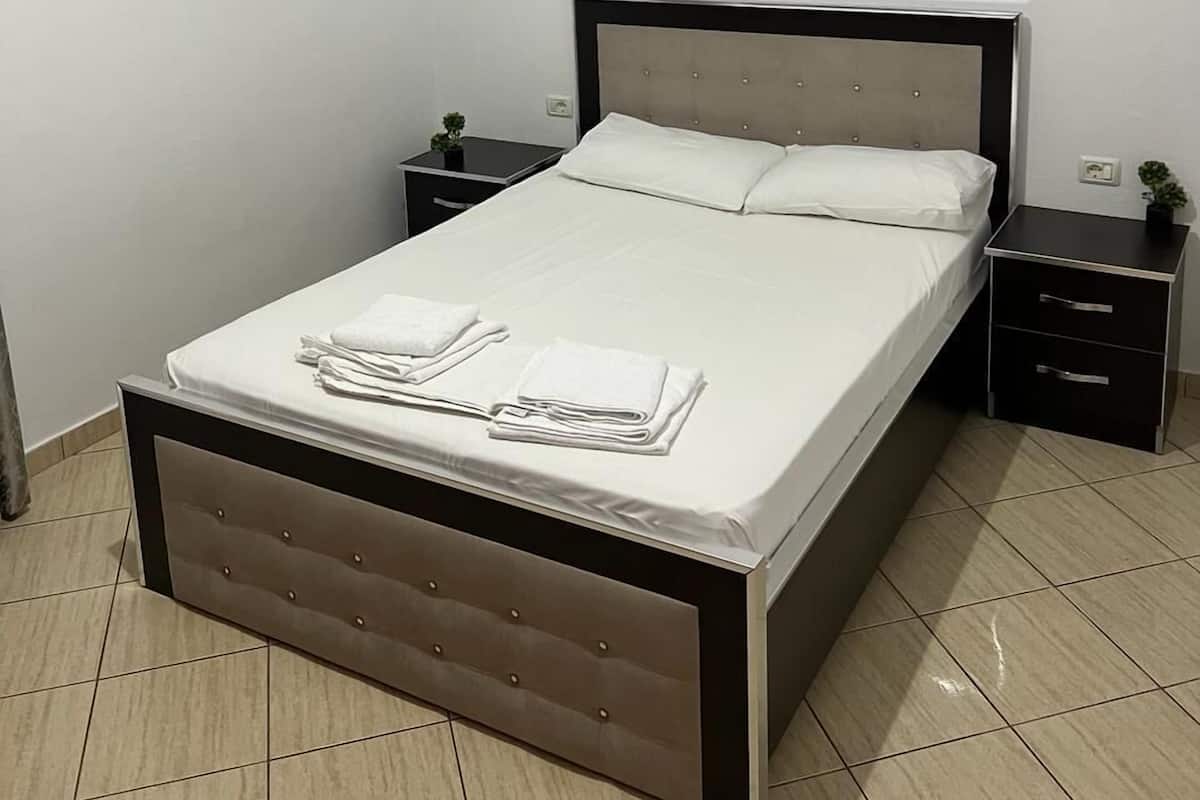 Una cama bien hecha con sábanas blancas y una toalla doblada en el reposapiés, una mesita de noche con una pequeña planta y un tocador con cajones.