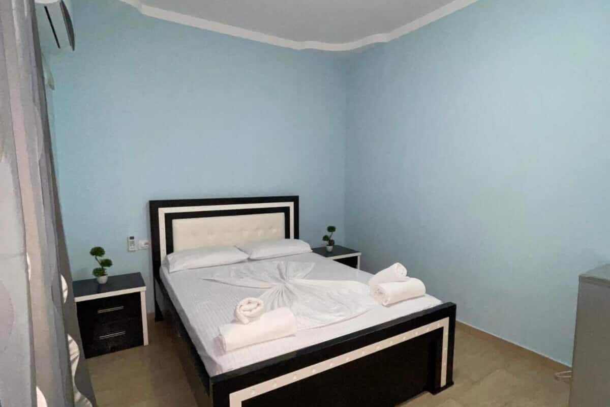Una cama bien hecha con sábanas blancas y una decoración de toallas, una mesita de noche con una planta y una pequeña mesita con una lámpara.