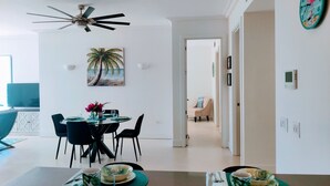 Una sala de estar moderna con comedor, que incluye un ventilador de techo, un reloj montado en la pared y un cuadro de una playa.