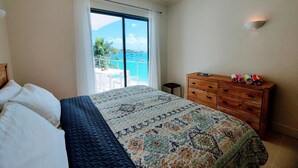 Un dormitorio con una cama, un aparador de madera, un ventilador de techo y vista al mar a través de un ventanal grande.
