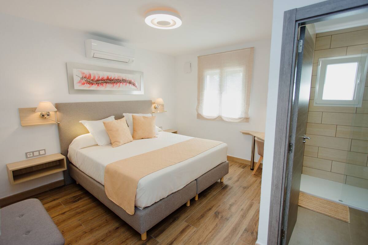 Une chambre à coucher comprenant un lit, des tables de chevet, une commode, une fenêtre avec des rideaux et une porte donnant sur une salle de bain.