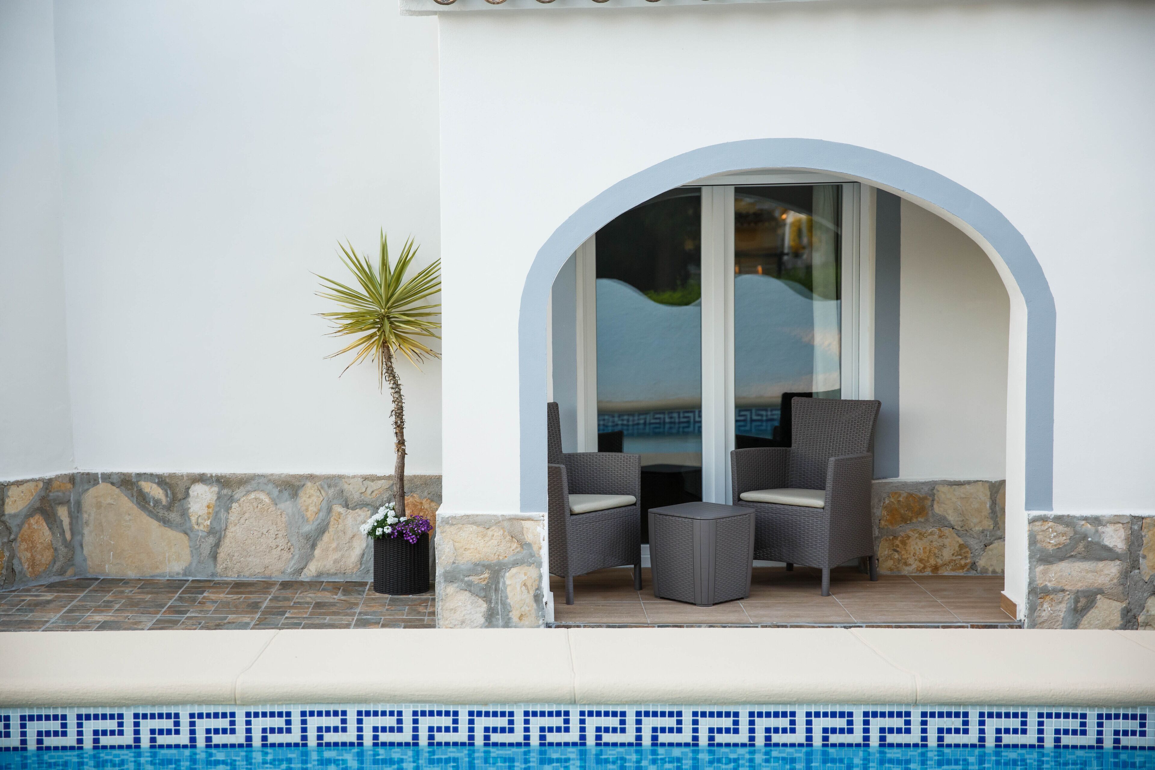 A poolside area with a stone wall, a small potted plant, and two wicker chairs with cushions.
