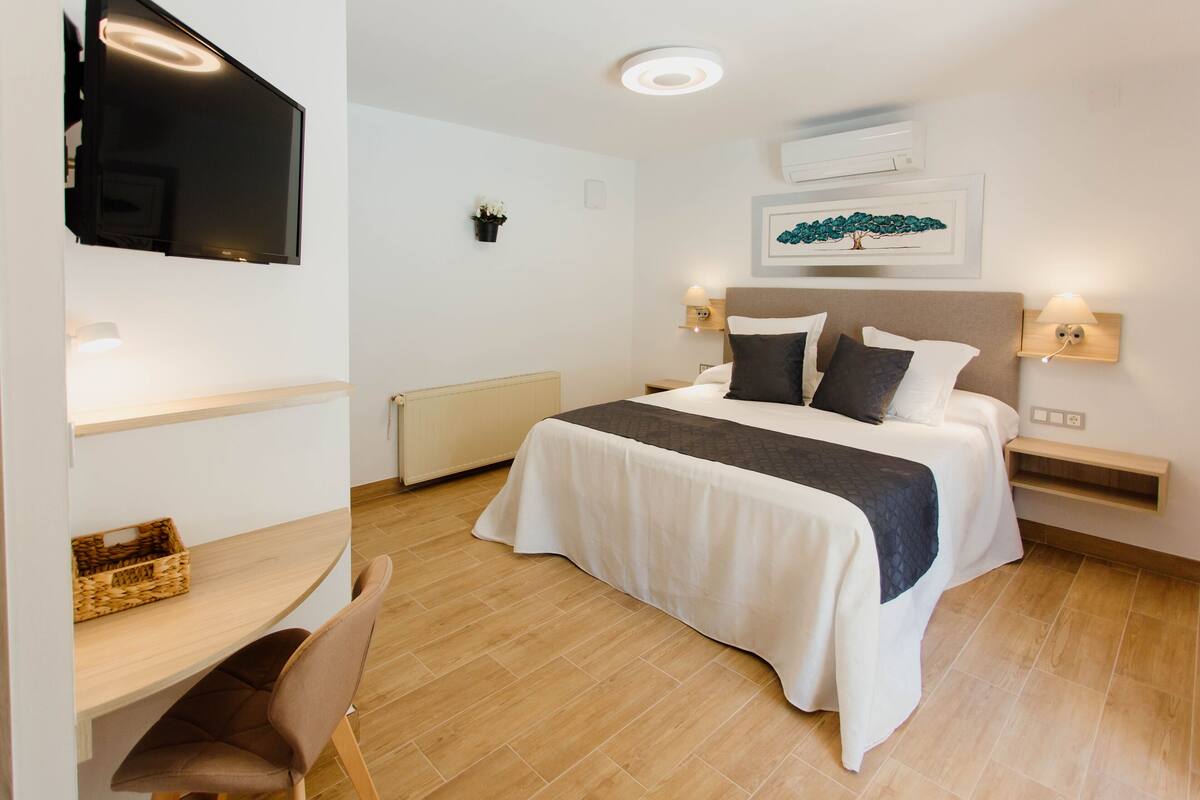 Une chambre d’hôtel comprenant un lit, des tables de chevet, un téléviseur fixé au mur, un bureau avec une chaise et une petite plante en pot.