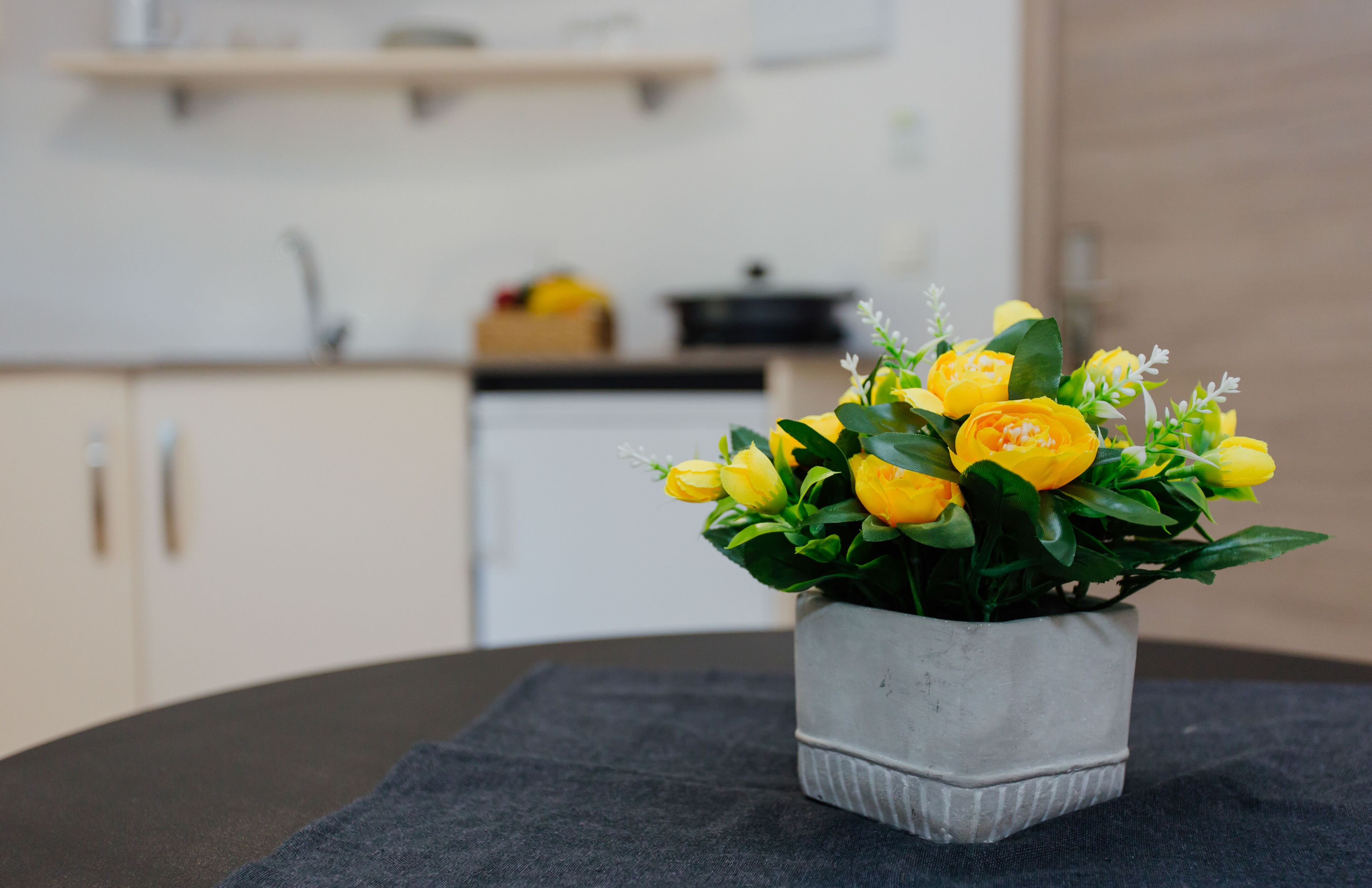 Una planta en maceta con flores amarillas sobre una mesa en una habitación con una cocina al fondo.