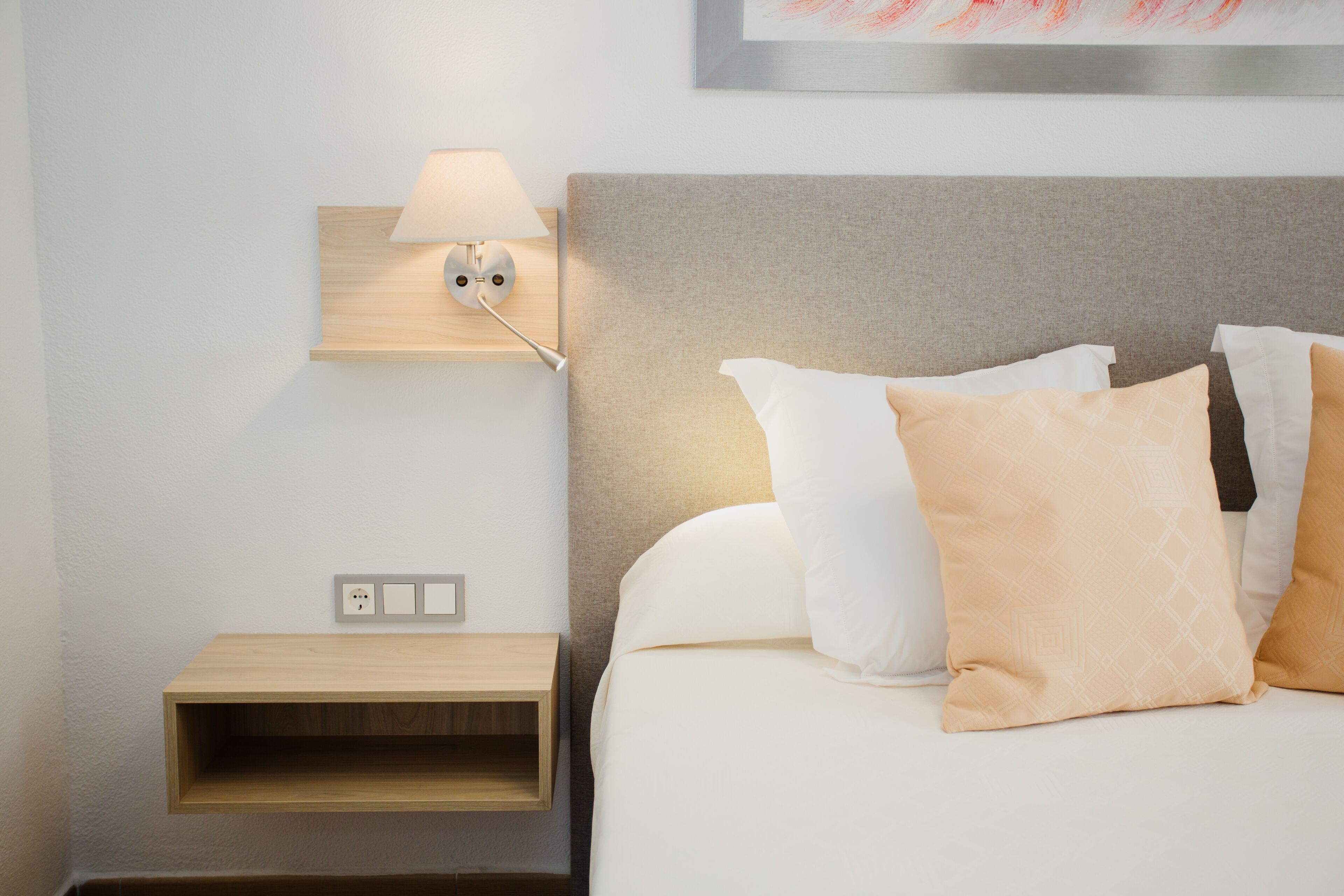 Habitación de hotel ordenada con cama, mesita de noche con lámpara y estante en la pared.