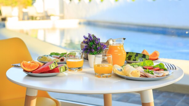 Una mesa con una variedad de alimentos para desayuno, incluyendo frutas, verduras y bebidas, cerca de una piscina con una valla blanca y vegetación al fondo.