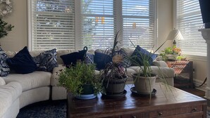 Ein Wohnzimmer mit einem beigefarbenen Sofa, blauen und weißen Kissen und mehreren Blumentöpfen auf einem Holztisch.