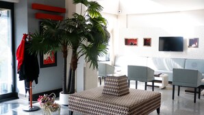 A modern living room with a large potted plant, a checkered ottoman, a grey sofa, and a flat-screen TV mounted on the wall. - Hotel La Bella Soma (Soma)