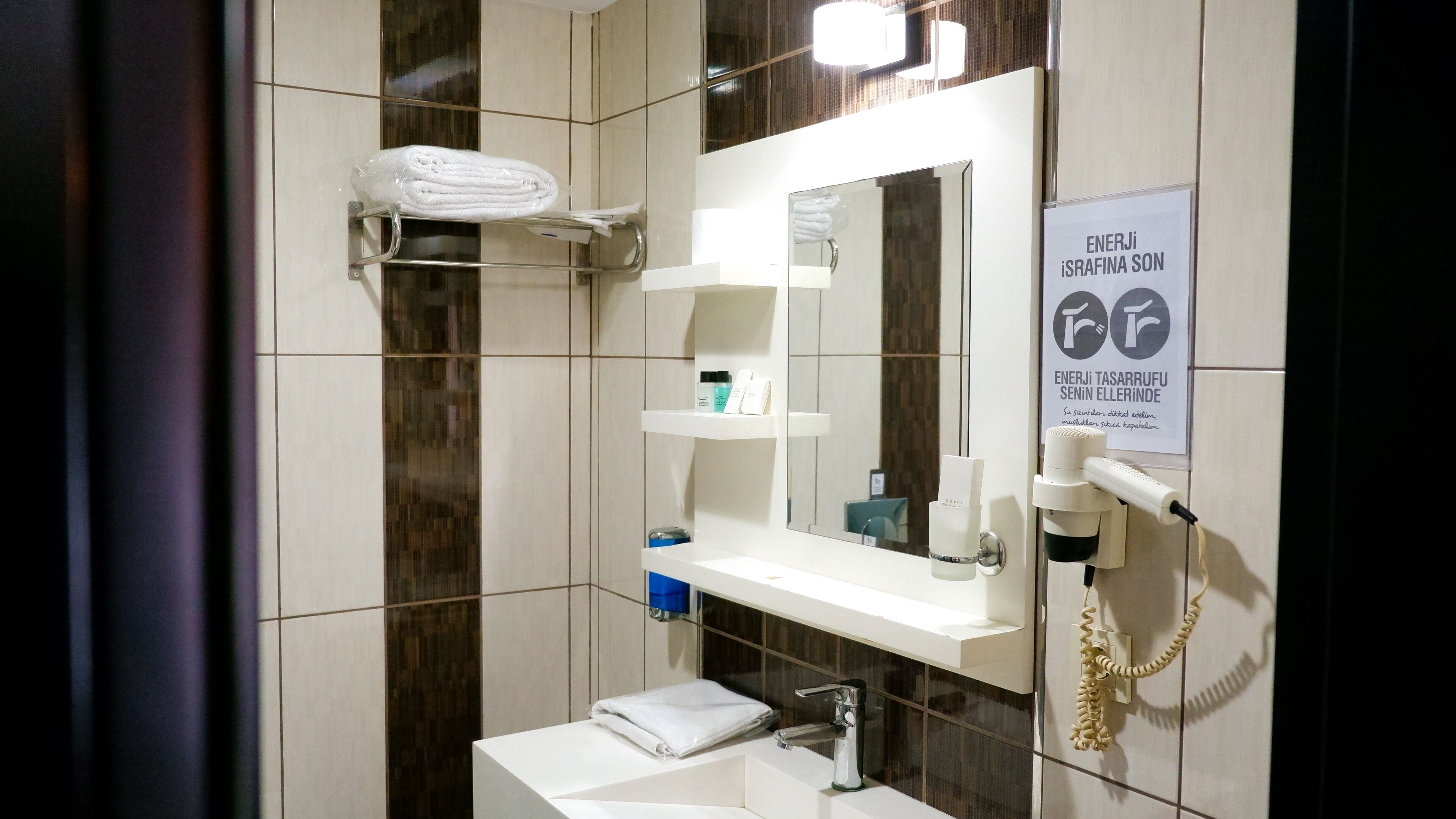 Un baño moderno con un lavamanos blanco, espejo y toallero. Hay un cartel en la pared y un secador de pelo montado en la pared.