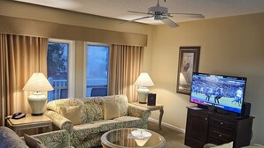 Ein Wohnzimmer mit floralen Sofas, einem Glastisch und einem Fernseher, der ein Sportereignis zeigt.
