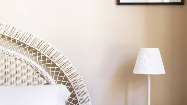 Une chaise en osier avec un coussin blanc, un lampadaire blanc et un tableau encadré au mur.