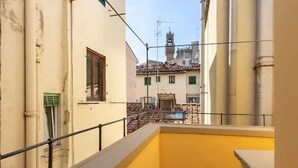 Un balcon avec un mur jaune et une rambarde en métal. Au fond, on aperçoit des bâtiments avec un clocher et un toit recouvert de tuiles.
