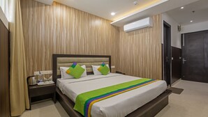 Een hotelkamer met een wand van houten panelen, een bed met witte beddengoed en kleurrijke kussens, een nachtkastje met waterkoker en fles water, en een airconditioner aan de muur.