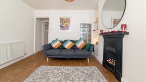 A living room with a grey sofa, a patterned rug, a black fireplace, a large mirror, and a door leading to another room.