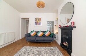 A living room with a grey sofa, a patterned rug, a black fireplace, a large mirror, and a door leading to another room.