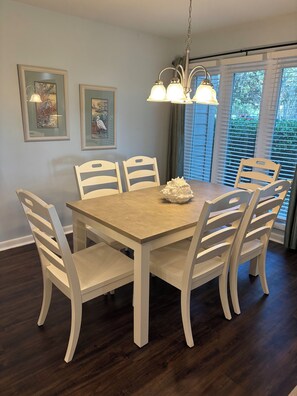 A dining area with a wooden table and white chairs, framed artwork on the wall, and a window with blinds.