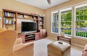 A living room with a television, bookshelves, a sofa, and a window with a view of trees. - Bear Trap Dunes Resort Bethany Beach 4BR, 3.5 Bath (Ocean View)
