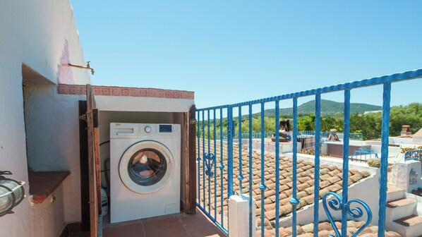 A laundry room with a washing machine, a blue metal gate, and a view of a tiled roof and greenery.