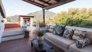 A covered patio with a sofa, a decorative vase, and a view of mountains. - Panoramic Attic with Pool | Near Costa Smeralda (Olbia)