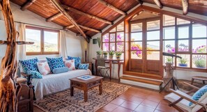 A cozy room with a wooden ceiling, a bed with patterned pillows, a small table, and a view of purple flowers outside.