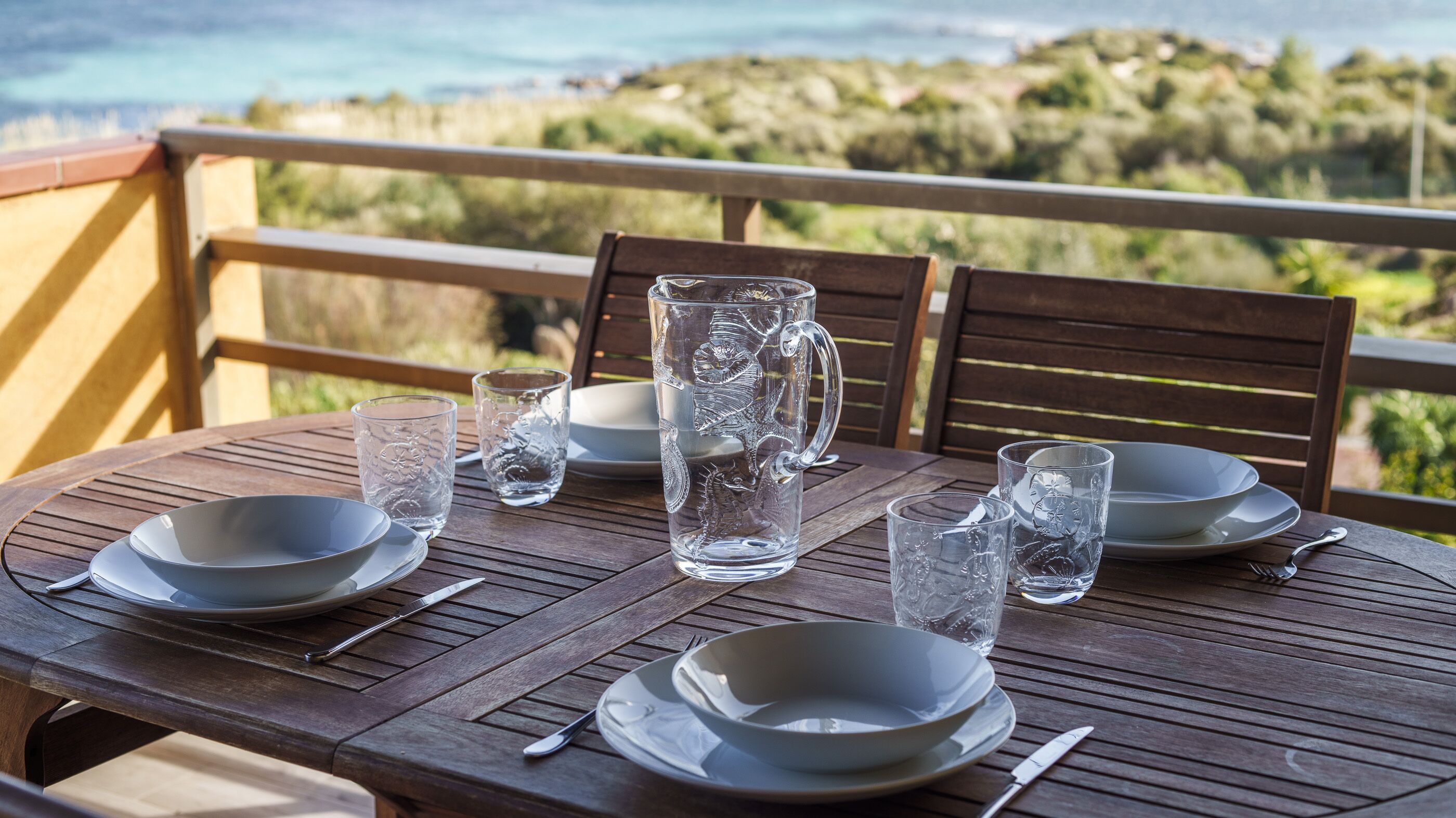A wooden table set for dining with clear glassware and plates, overlooking a scenic landscape.