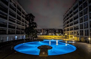 Une piscine nocturne avec une fontaine centrale, entourée d’immeubles à plusieurs étages.