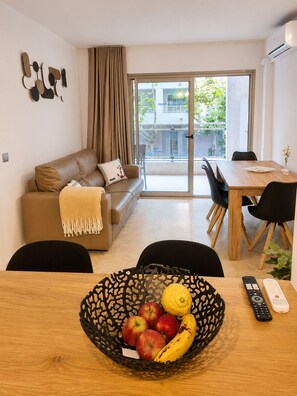 A modern living room with a sofa, a dining table with chairs, a balcony with a sliding door, and a fruit basket on the table.