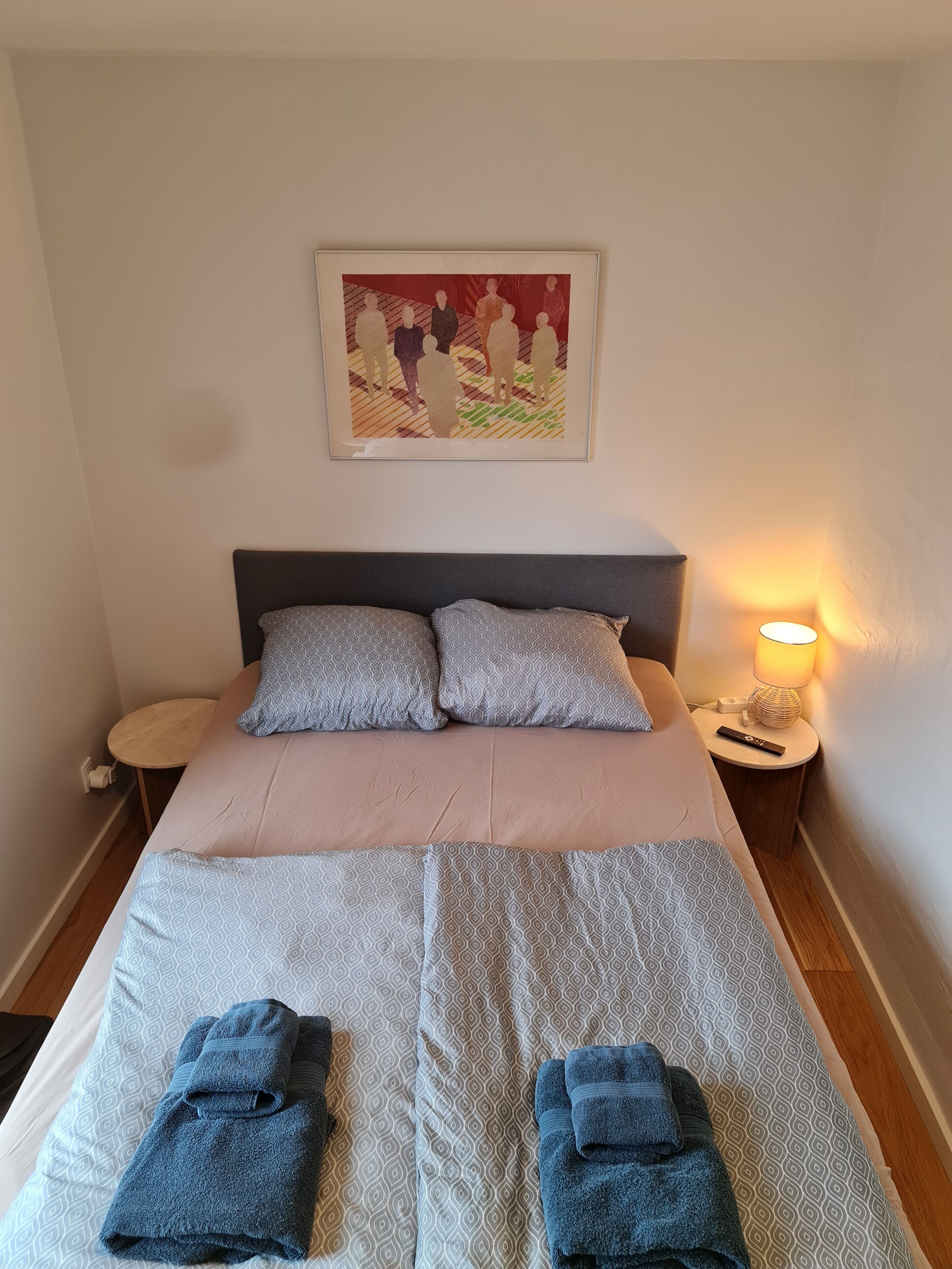 Un llit ben fet amb roba de llit estampada, dos coixins, una làmpada de nit i un quadre al mur.