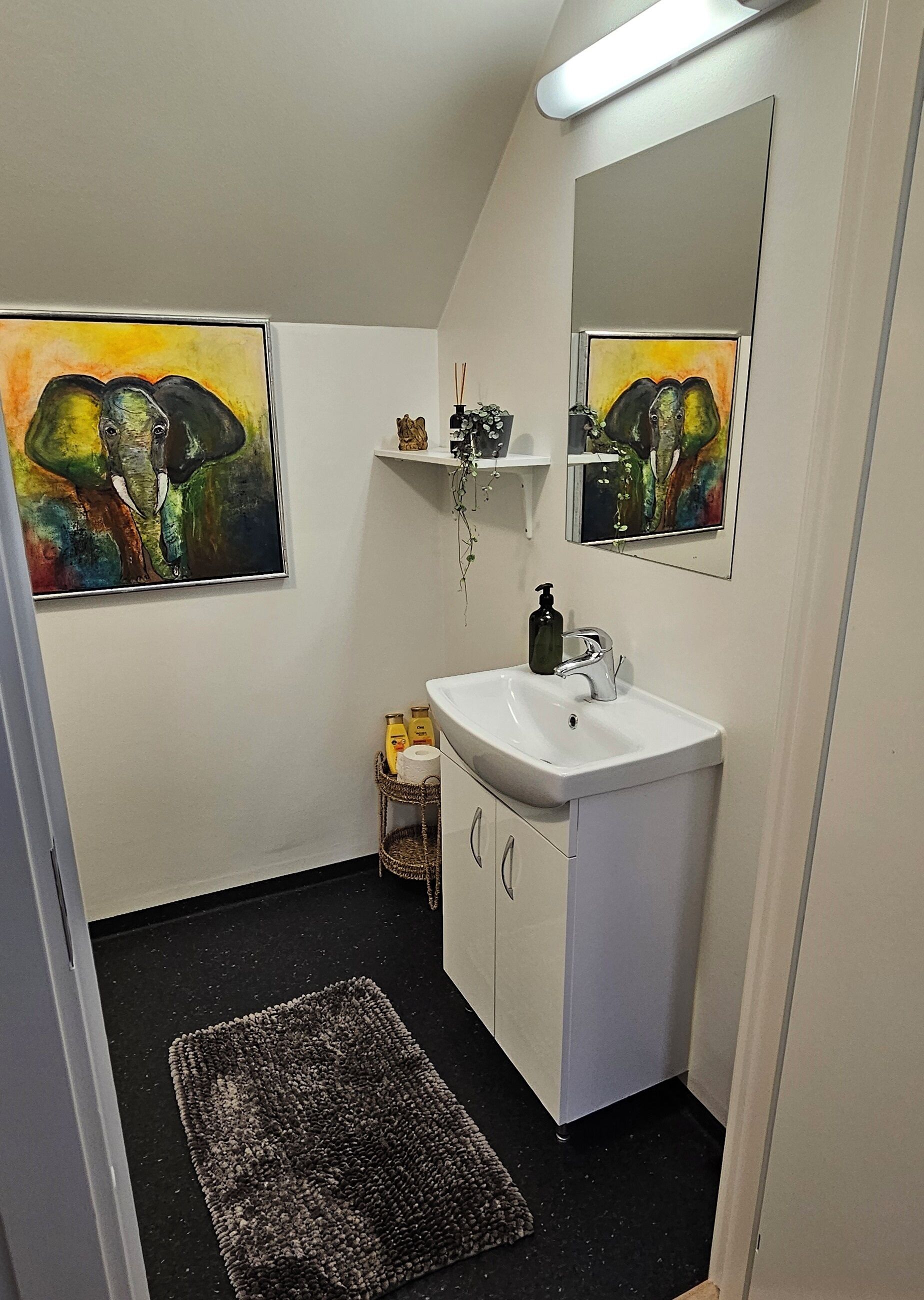 Un baño con un lavabo blanco, un espejo y dos cuadros de elefantes. Hay estantes con objetos decorativos y una pequeña cesta con artículos de aseo.