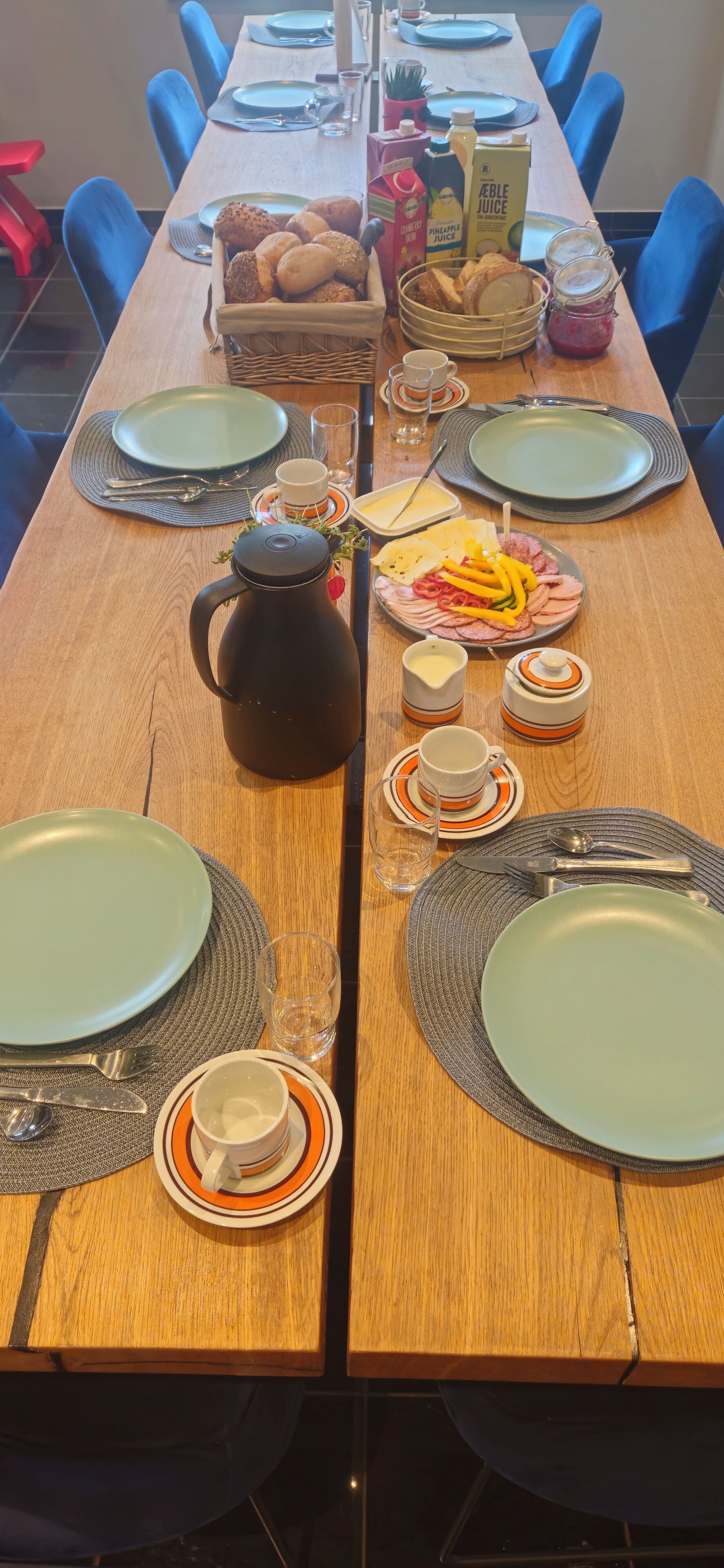 Una mesa de comedor dispuesta con platos, cubiertos, una cafetera y una cesta de pan.