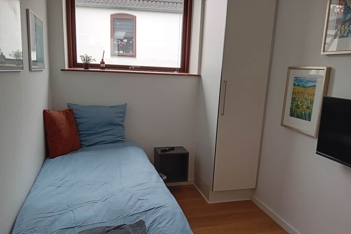 A small bedroom with a single bed, a window, a wardrobe, and a TV mounted on the wall.