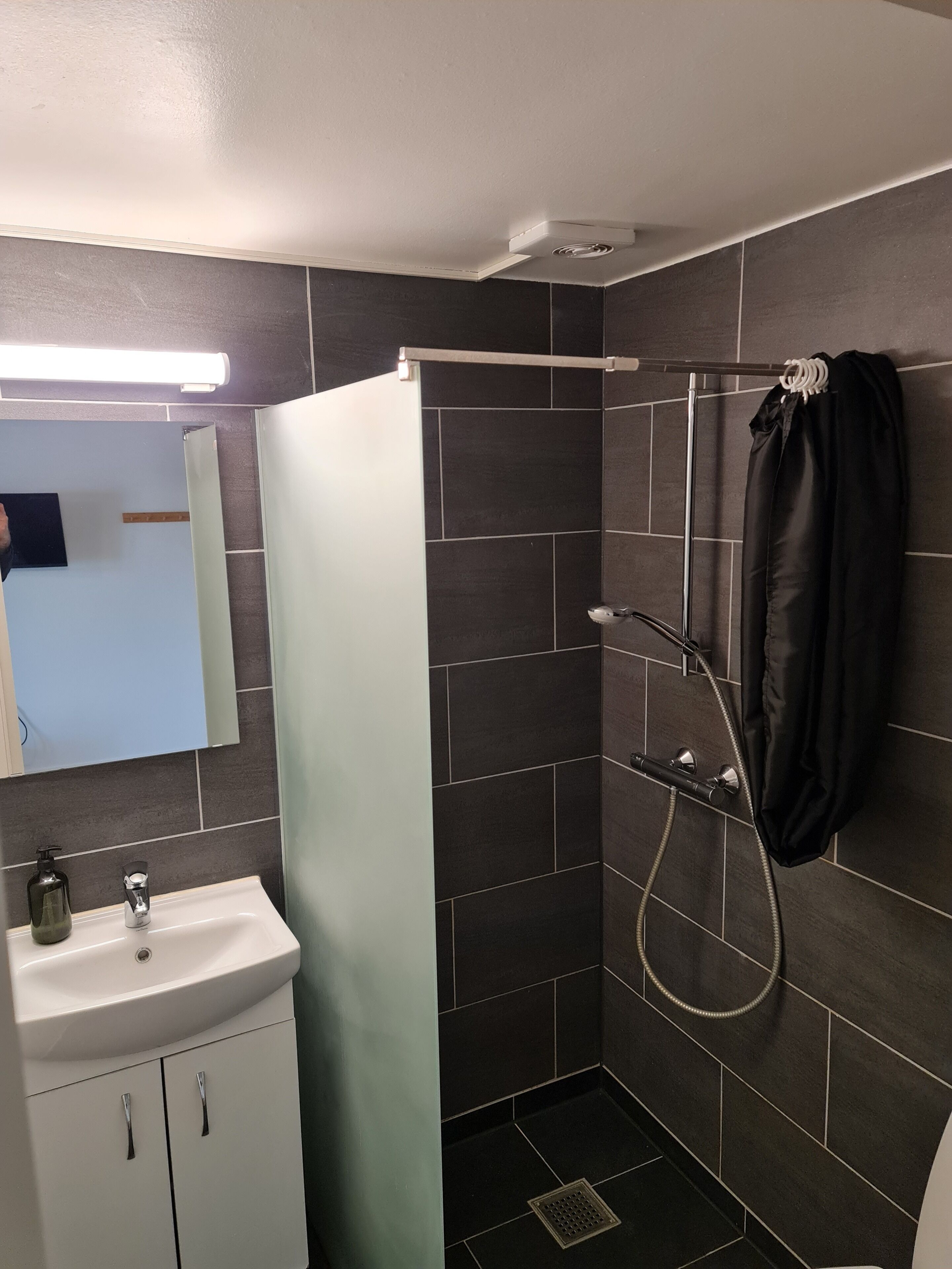 A bathroom with a white sink, a mirror, a shower with a black towel, and dark tiled walls.