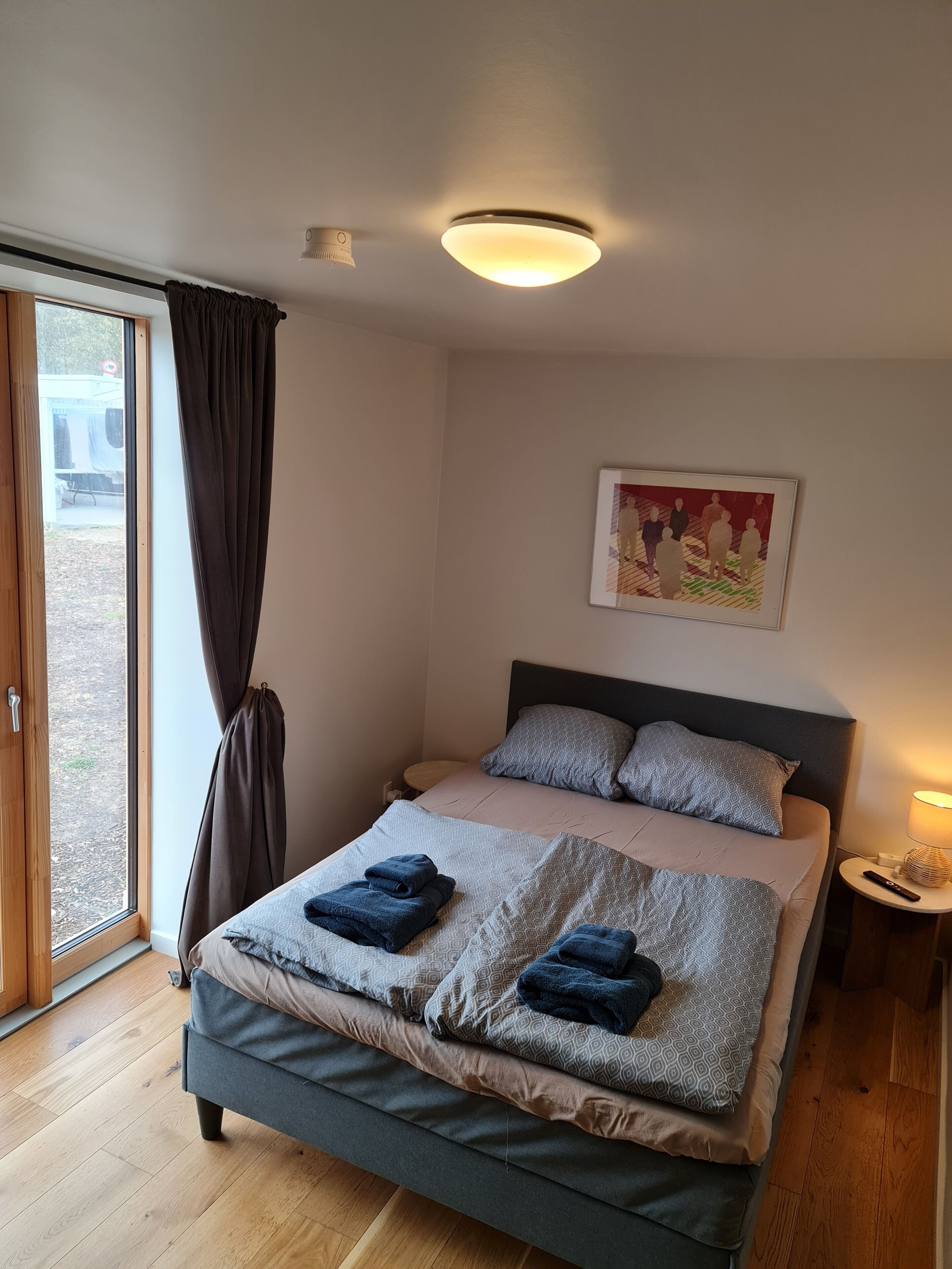 Une chambre à coucher avec un lit, une tête de lit, une table de chevet avec une lampe, une fenêtre avec des rideaux et un tableau au mur.
