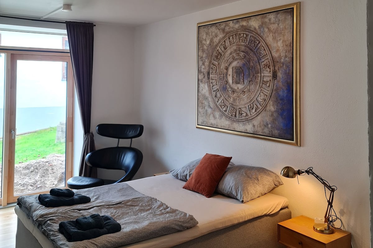A bedroom with a bed, a chair, a nightstand, and a large wall painting.