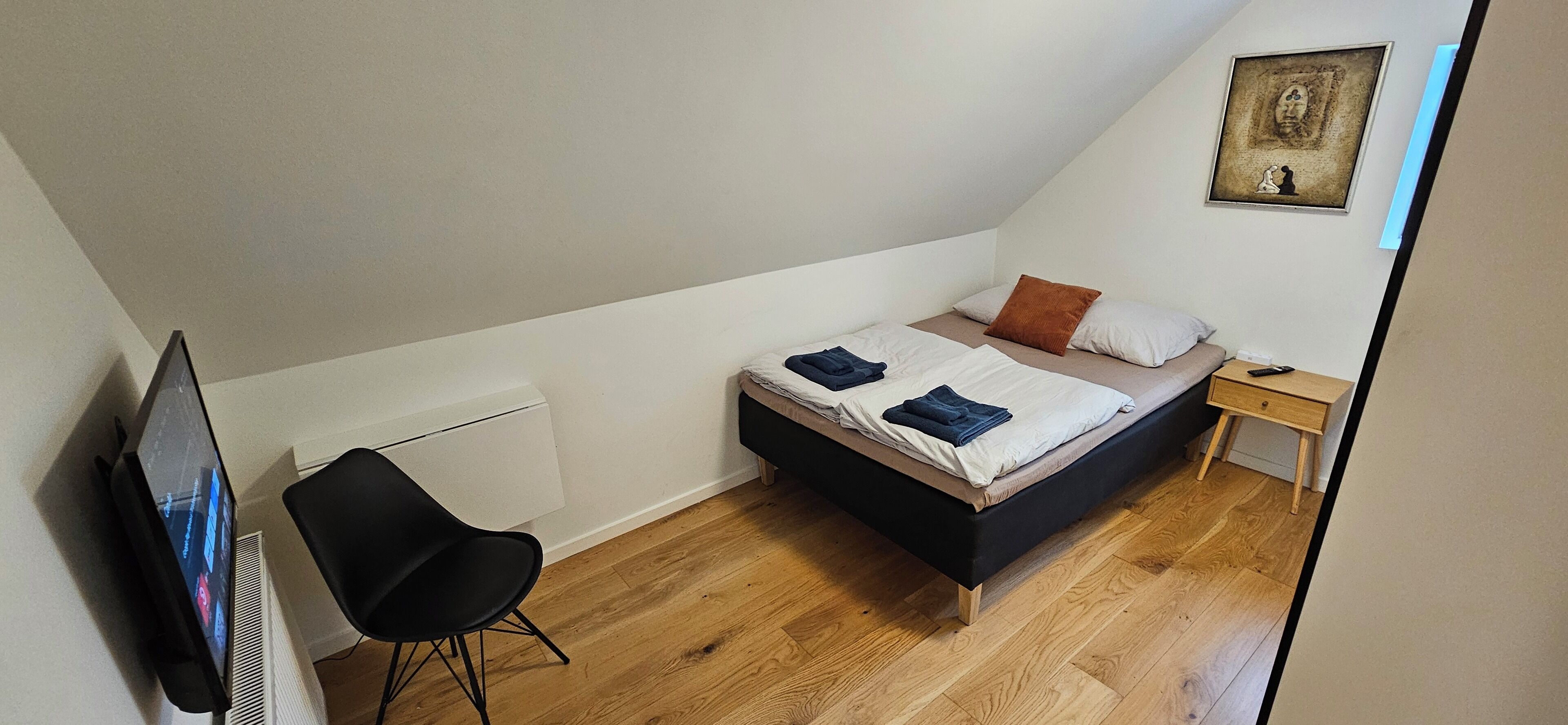 A small, modern bedroom with a bed, a chair, a bedside table, and a TV.