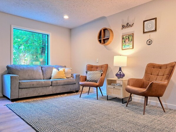 A living room with a grey sofa, two armchairs, a small table with a lamp, and a large window with a view of trees.