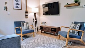 Una sala de estar con dos sillones azules, una mesa de centro de madera, un tapete estampado, un televisor montado en la pared y un estante con objetos decorativos.