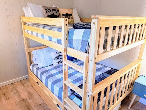 A bunk bed with a checkered blue and white bedding, a pillow with "TO FISH" text, and a small blue side table.