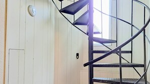 Un escalier en colimaçon avec des rampes en métal, situé dans une pièce aux murs en bois peints en blanc et dotée d’une grille d’aération dans le plancher.