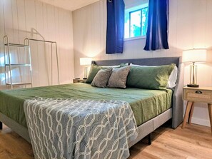 Un lit bien fait, recouvert d’une couverture verte et d’un plaid à motifs, une table de chevet avec une lampe, et une fenêtre ornée de rideaux bleus.