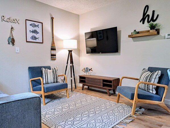 A modern living room with two blue armchairs, a wooden coffee table, a patterned rug, a TV mounted on the wall, and a small shelf with decorative items.