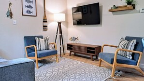 A modern living room with two blue armchairs, a wooden coffee table, a patterned rug, a TV mounted on the wall, and a small shelf with decorative items. - Timber Point - Newly Renovated Indian Point Cabin (Branson)