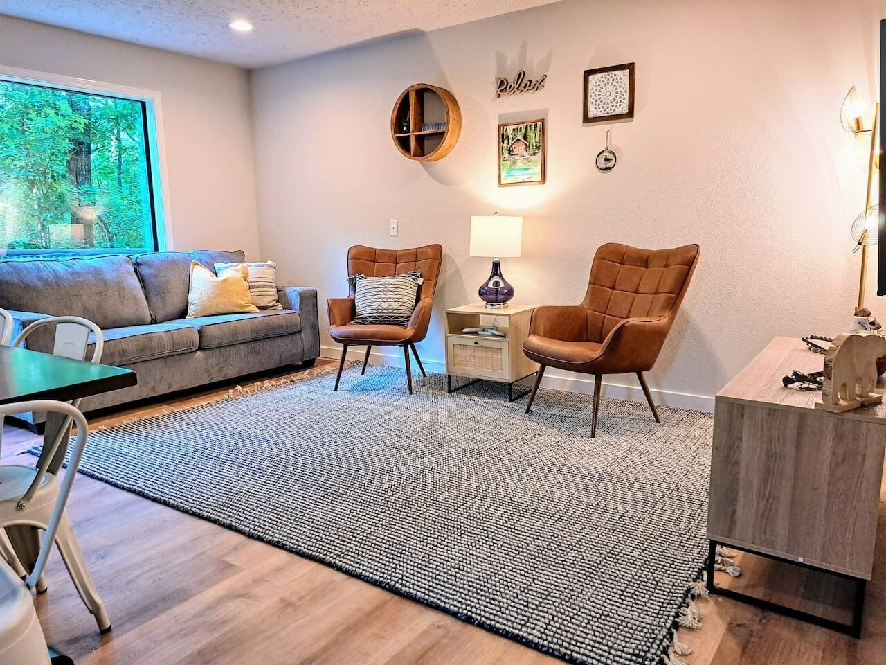 Un salon moderne avec un canapé gris, deux fauteuils en cuir marron, une table d’appoint en bois avec une lampe, et une grande fenêtre donnant sur des arbres.