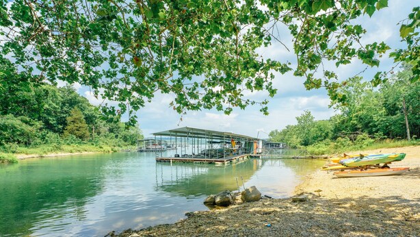 A riverside dock with a covered structure, calm water, and a gravel path leading to a boat. - Tribesman Resort 12 - Renovated Condo, Lake Access, Slip Rental, Great Location! (Branson)