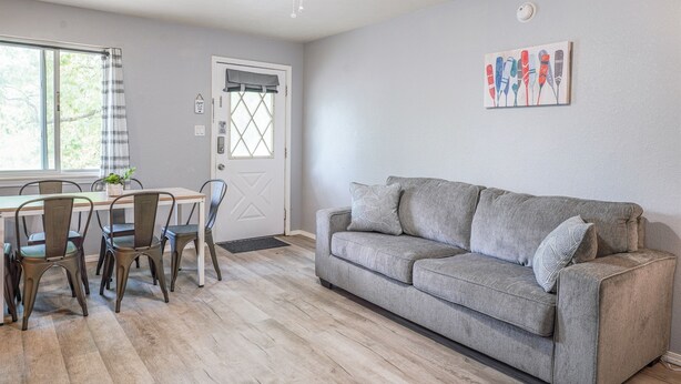 Een woonkamer met een grijze bank, een eettafel met stoelen, een plafondventilator en een raam met gordijnen.