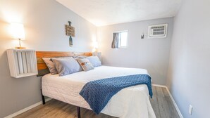 Een slaapkamer met een houten hoofdbord, een witte spreideken en een blauwe plaid. Er is een wandairconditioner en een klein raam met gordijnen.