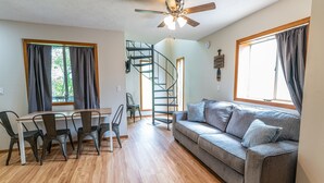 A modern living room with a wooden floor, a gray sofa, a dining table with chairs, a ceiling fan, and a spiral staircase. - Tribesman Resort #22 - Renovated Cabin, Minutes to Silver Dollar, Great Location (Branson)