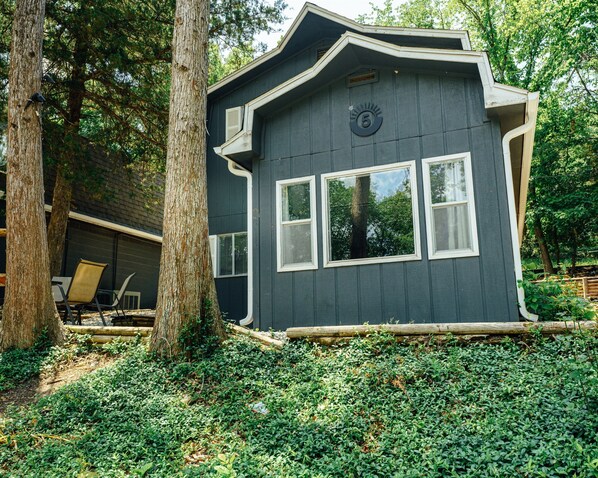A small, dark-colored cabin with three windows, surrounded by trees and a garden.