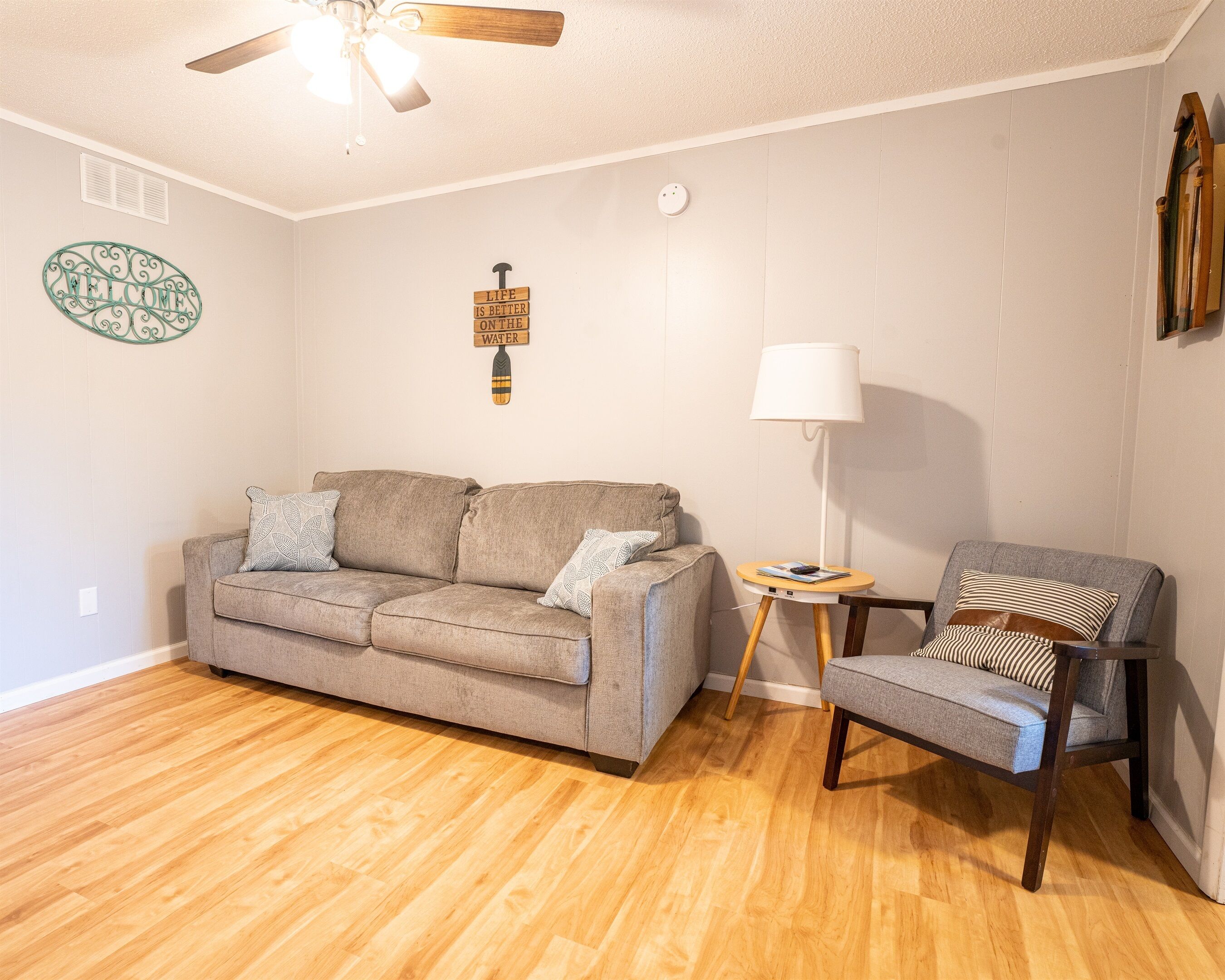 A living room with a wooden floor, a grey sofa, a grey armchair, a small table with a lamp, and a ceiling fan.